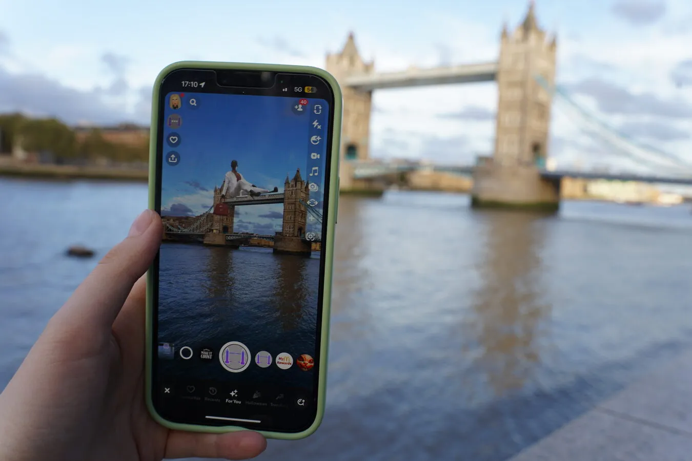 A phone held up at the riverside showing a Snapchat lens with a mannequin world-locked on top of Tower Bridge, with the real bridge behind it