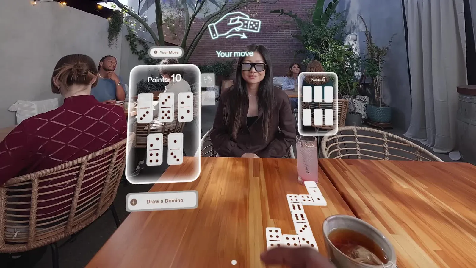 Point of view through Snap Specs at a cafe table, facing an opponent also wearing Specs, with a hand of virtual dominoes and a shared line of dominoes on the real table